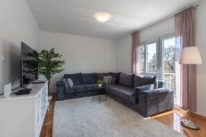 Uma sala de estar moderna com um sofá seccional cinza, um tapete branco, uma pequena mesa lateral com uma planta, uma TV de tela plana sobre um móvel branco e um abajur de chão.