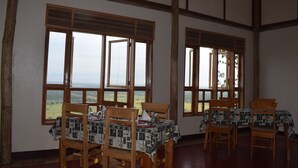A dining area with wooden chairs and tables, large windows with a view of the countryside, and a simple, rustic interior design. - Rift Valley Game Lodge (Rubirizi)