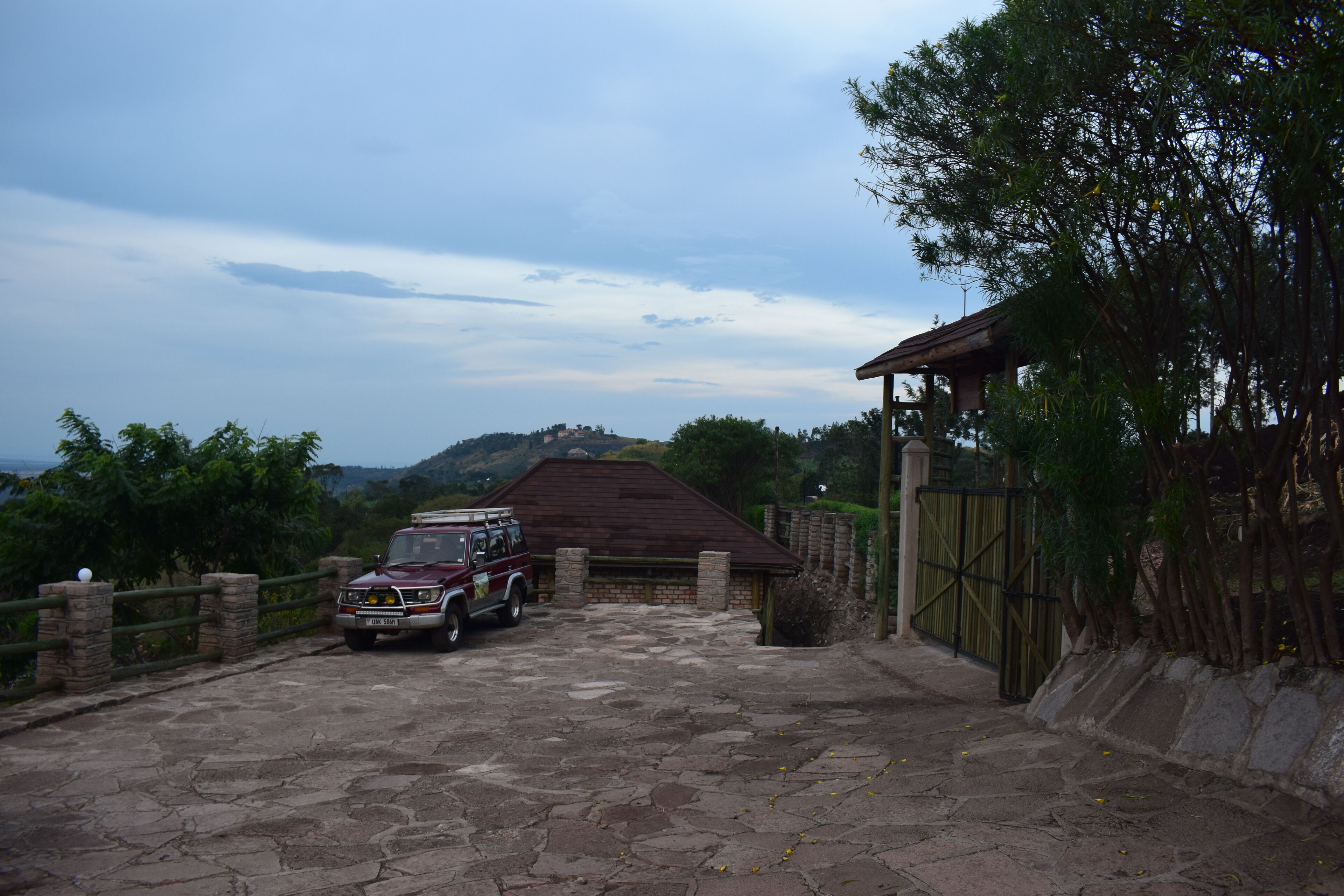 A stone-paved driveway leading to a wooden gate, a parked vehicle, and a building with a brown roof.