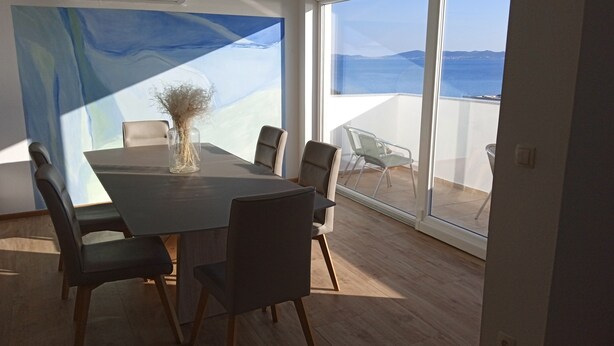 A modern dining room with a large table and chairs, a vase with dried plants, and a view of the sea through large windows.