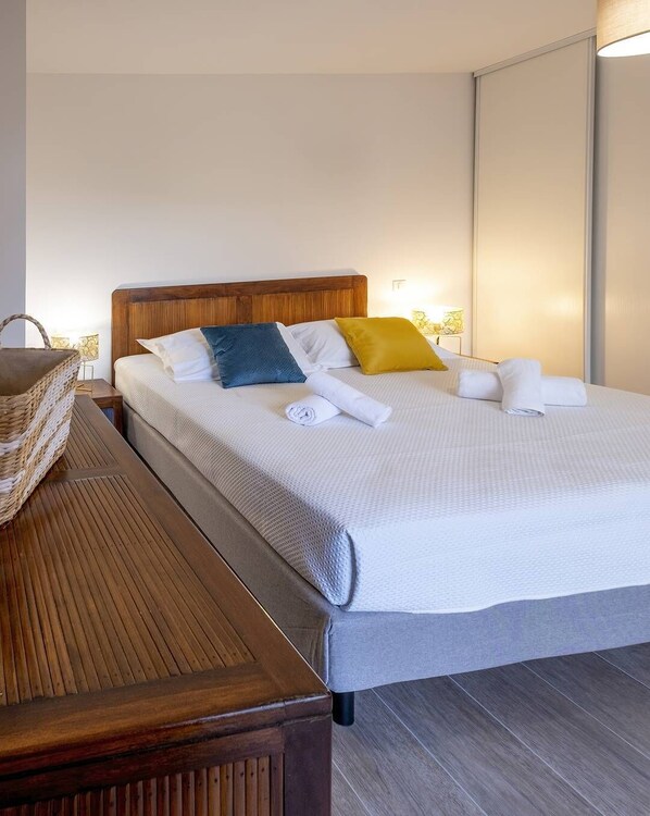 Un lit bien fait, avec une tête de lit en bois, une table de chevet surmontée d’un panier en osier et une lampe fixée au mur.