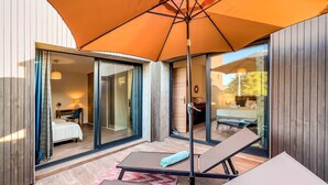 A modern hotel room with a glass door leading to a balcony, a bed, a desk, and a large orange umbrella providing shade. - Villa Renajolo. 6 pers, 3 min plage en voiture (STE LUCIE DE PORTO VECCHIO)