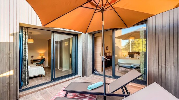 Una habitación de hotel moderna con una puerta de vidrio que da a un balcón, una cama, un escritorio y un paraguas naranja grande que proporciona sombra.