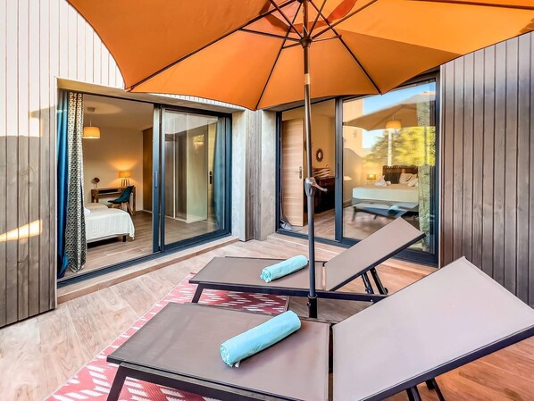 Una habitación de hotel moderna con una puerta de vidrio que da a un balcón, una cama, un escritorio y un paraguas naranja grande que proporciona sombra.