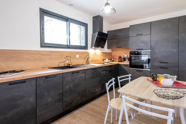 A modern kitchen with dark cabinets, a wooden countertop, a stainless steel sink, and a dining area with a table and chairs.