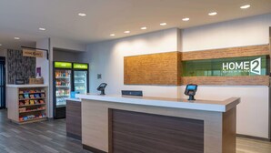 A hotel reception area with a reception desk, a self-service kiosk, a refrigerator with beverages, and a snack shelf. - Home2 Suites by Hilton Fredericksburg South (Fredericksburg)
