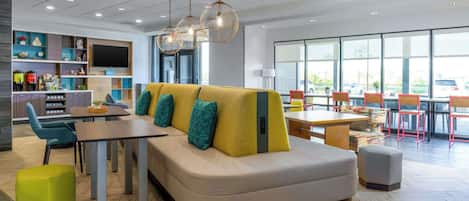 A modern hotel lobby with a seating area, a dining table, and a TV screen.