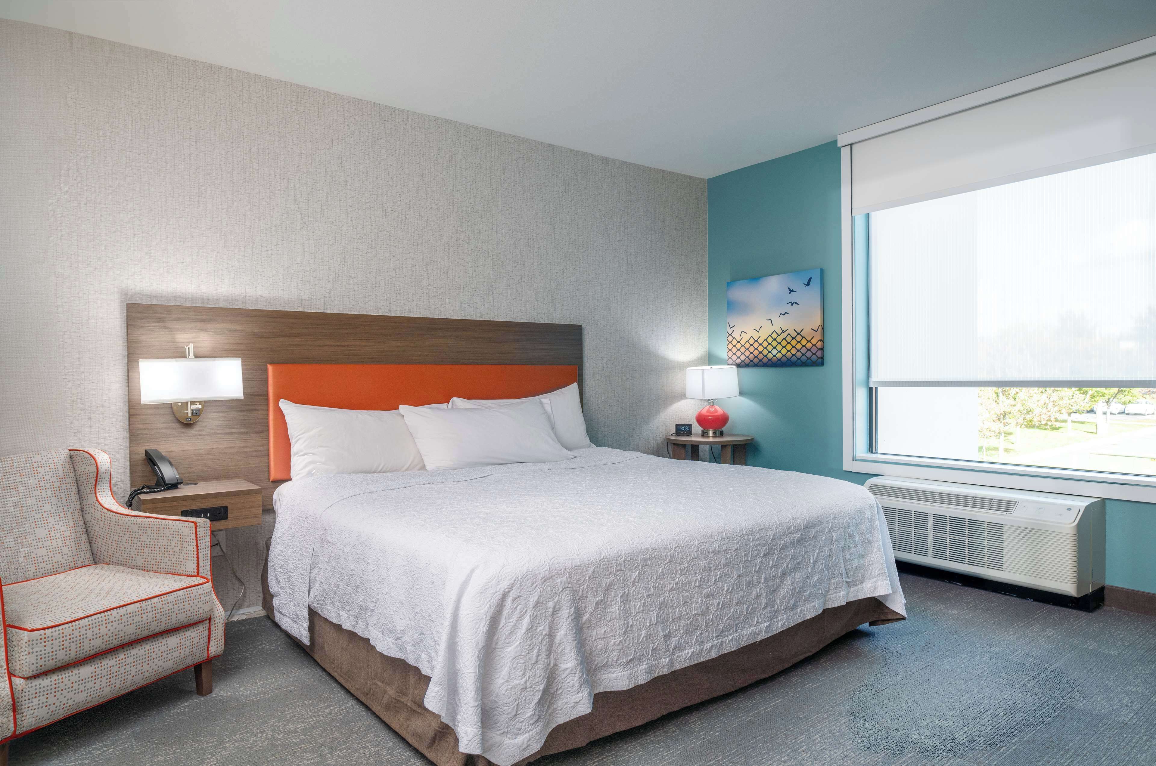 A hotel room with a bed, a chair, a nightstand, a lamp, a painting, and a window.
