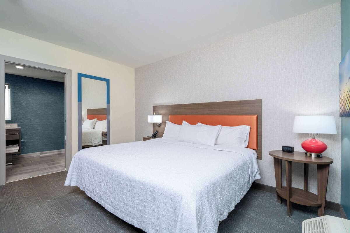 A hotel room with a large bed, a wooden headboard, a bedside table with a lamp, and a mirror on the wall.