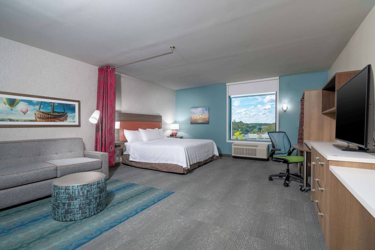 A hotel room with a bed, a desk with a computer, a sofa, a chair, and a TV.