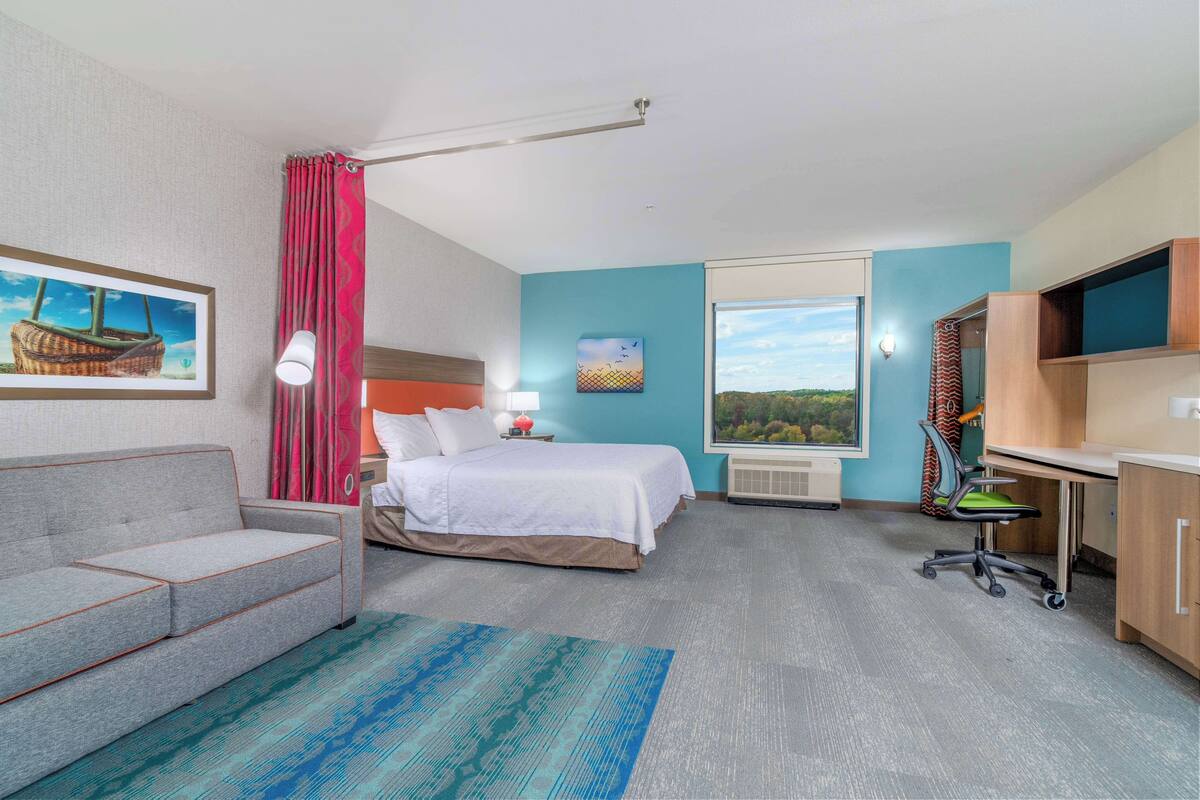 A hotel room with a bed, a desk, a chair, a sofa, a lamp, a painting, a window, and a television.