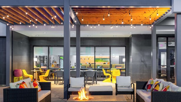 An outdoor seating area with wicker furniture, a fire pit, and a restaurant in the background. - Home2 Suites by Hilton Fredericksburg South (Fredericksburg)