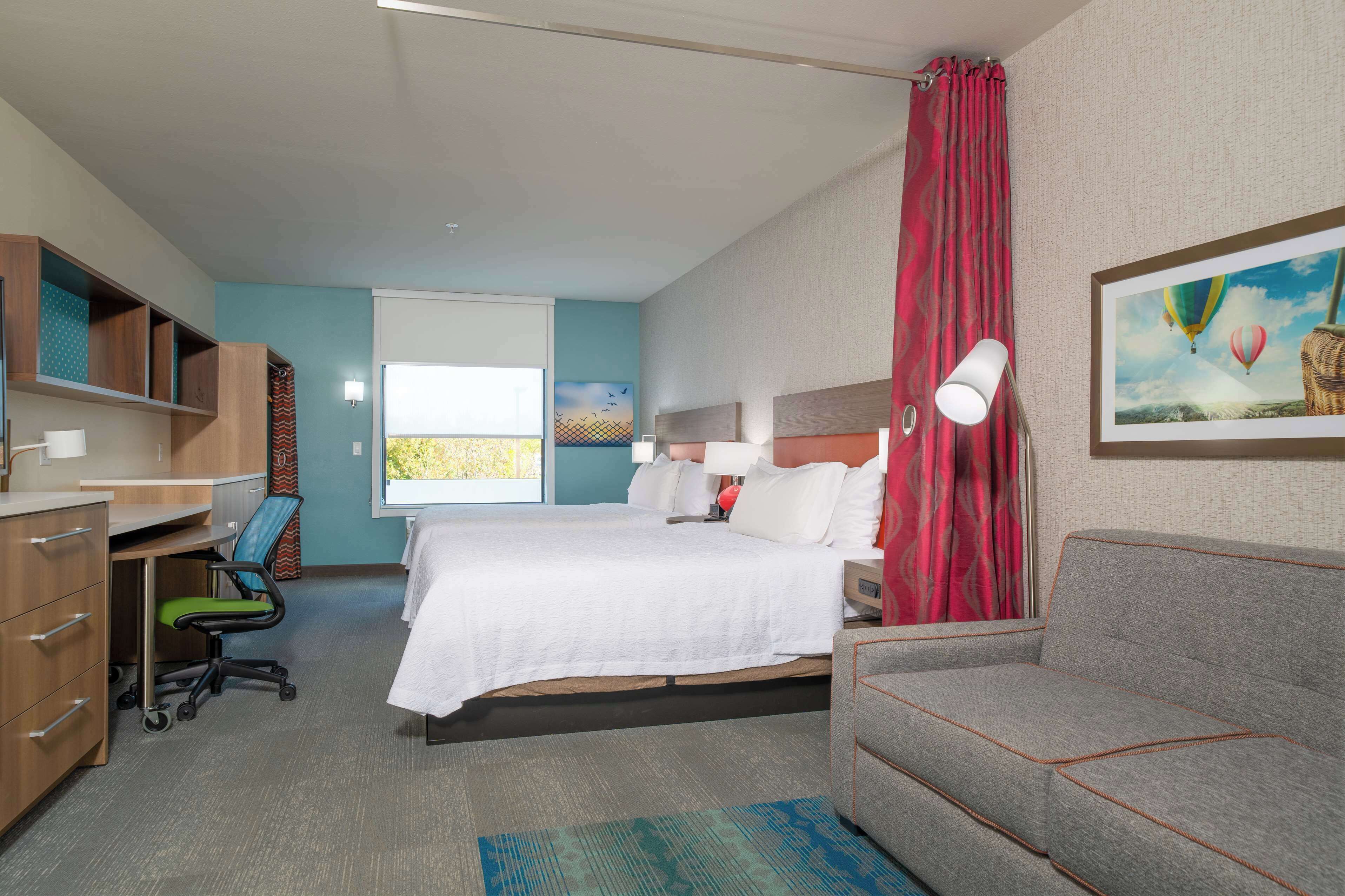 A hotel room with a bed, a desk, a chair, a sofa, a window with curtains, and a painting on the wall.