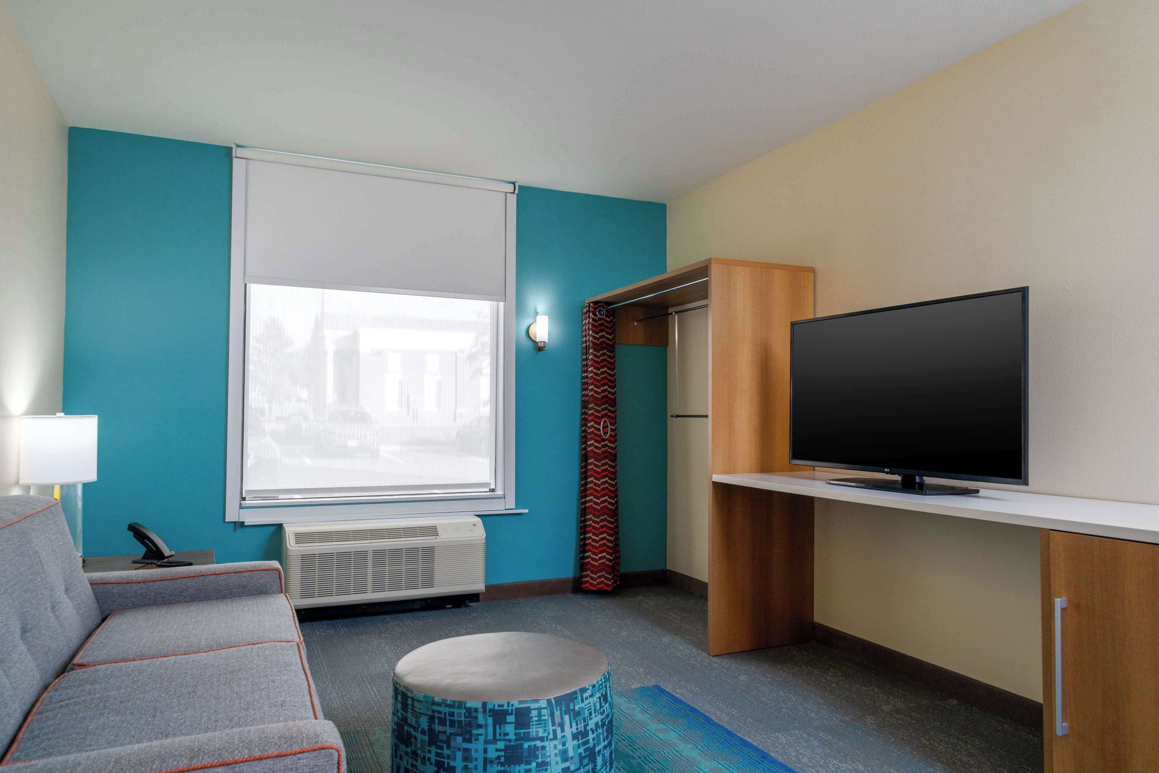 A modern hotel room with a grey sofa, a flat-screen TV, a window with blinds, and a patterned ottoman.