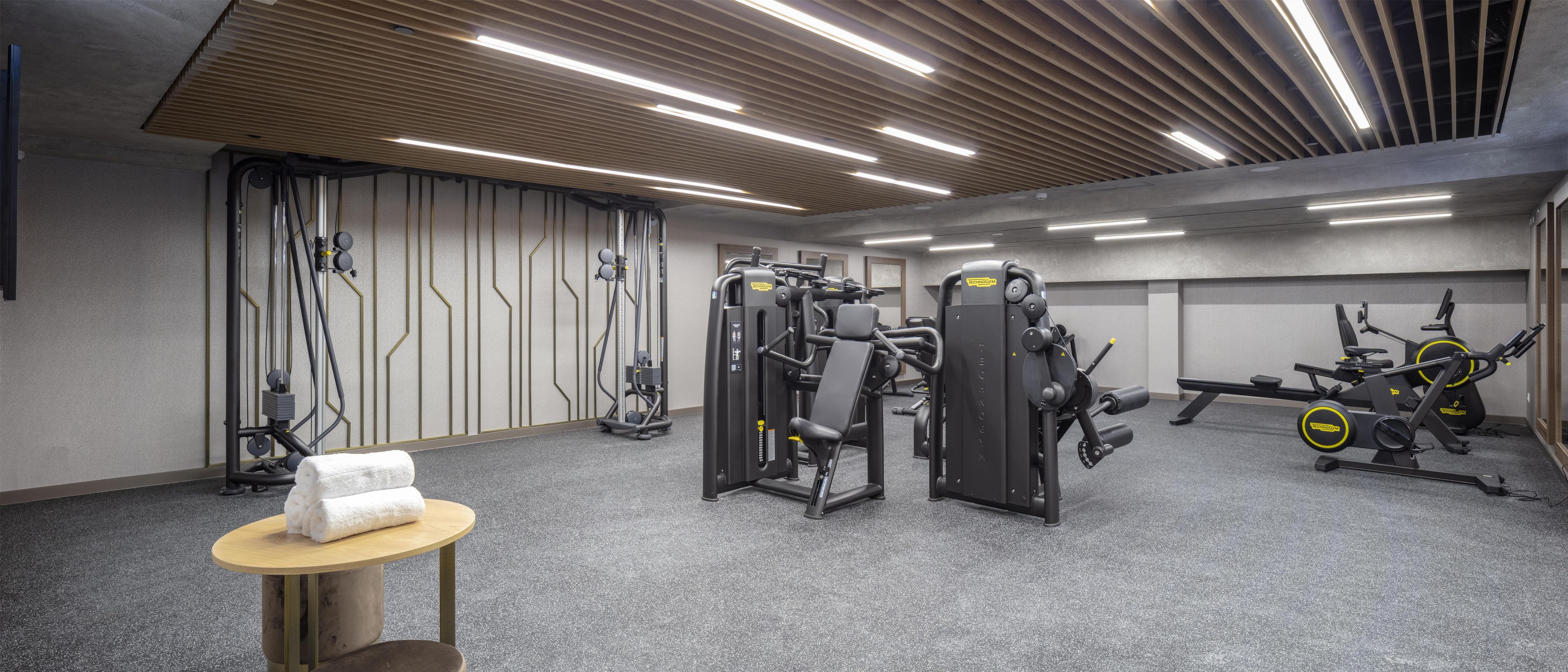 A gym with various exercise machines, a towel on a table, and a ceiling with linear lights.