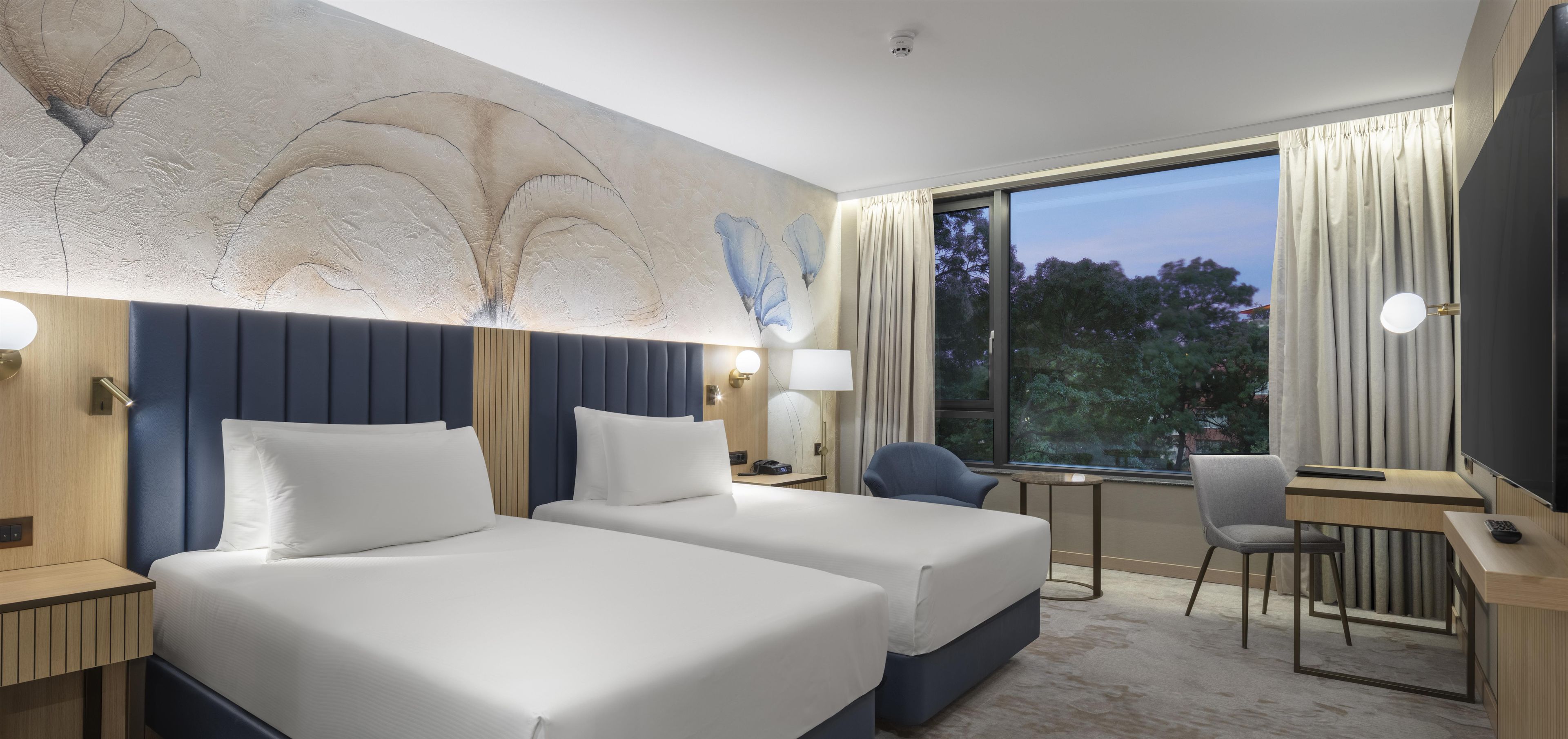 A hotel room with two beds, a desk, a chair, and a large window.