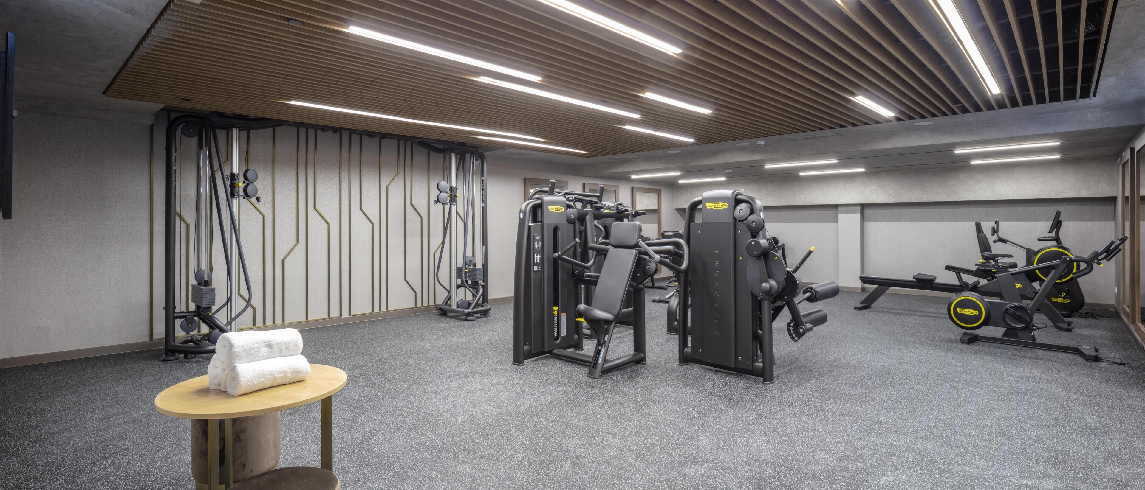 A gym with various exercise machines, a towel on a table, and a ceiling with linear lights.