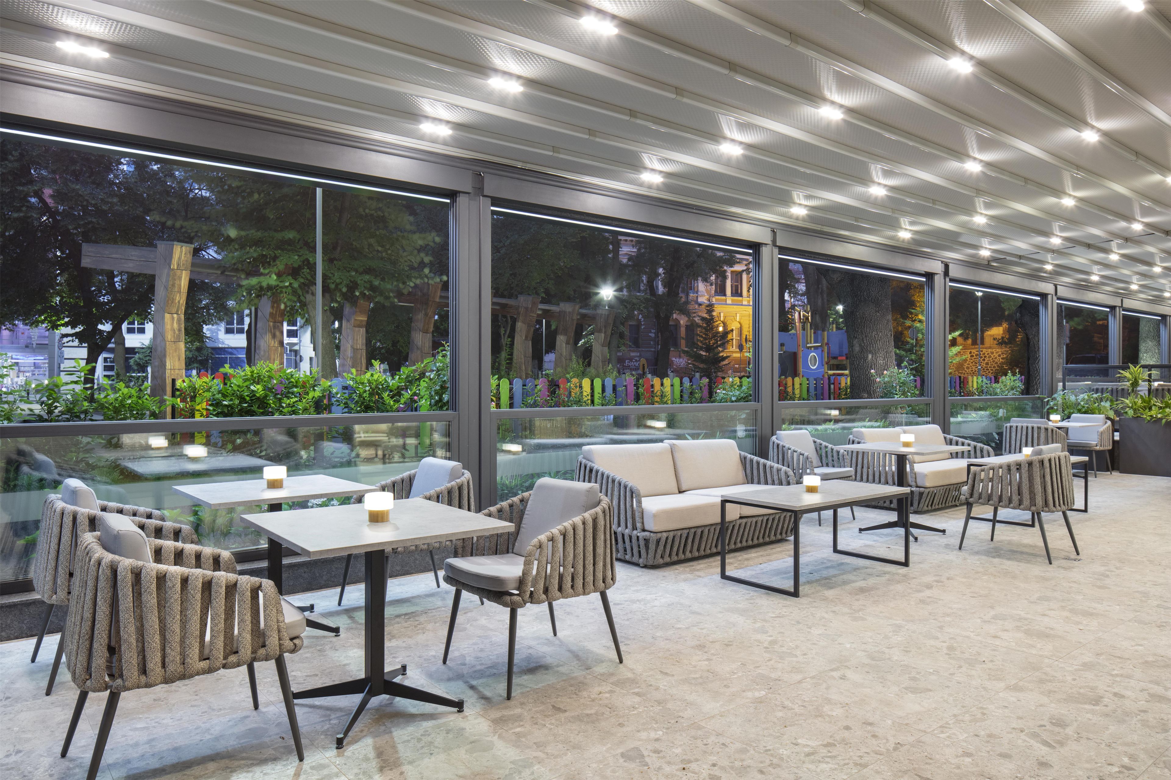 An outdoor seating area with tables and chairs, large windows, and a view of trees and buildings.