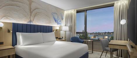 Habitación de hotel con una cama grande, un escritorio, una silla, una mesita y vista a la ciudad desde la ventana.