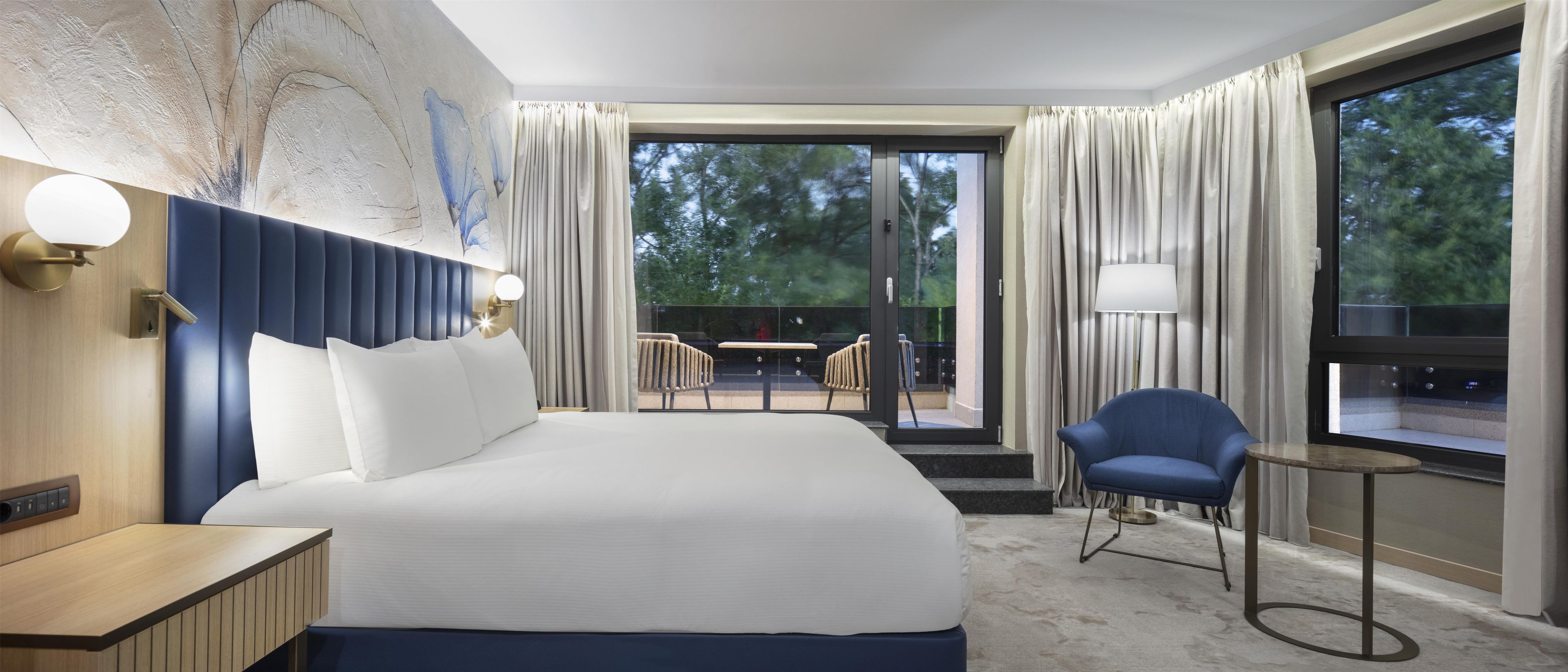 A hotel room with a large bed, a desk, a chair, and a view of the outdoors.