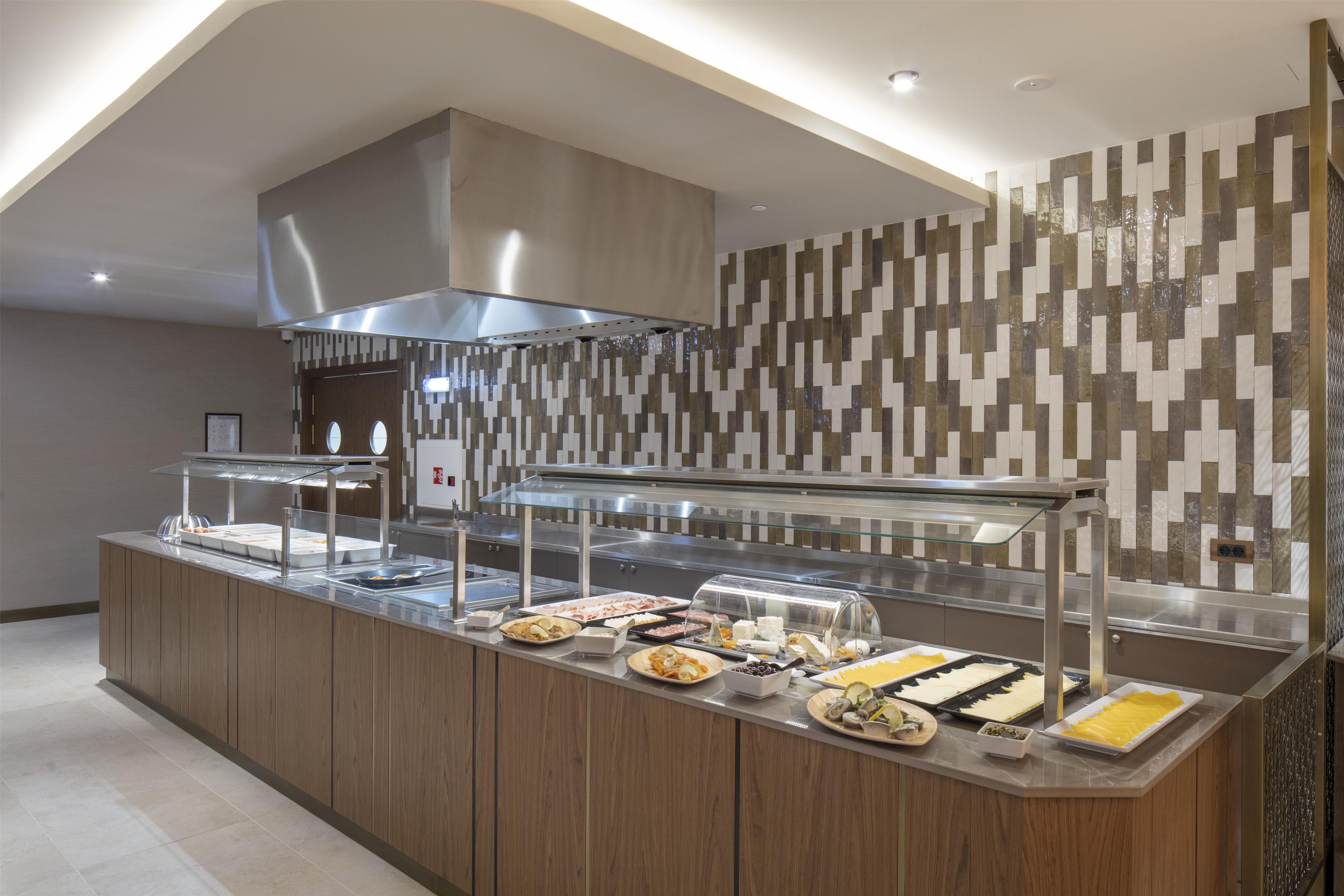 Zona de bufé con diversos alimentos, una campana extractora de acero inoxidable y una pared con diseño.