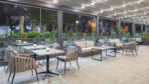 A modern outdoor seating area with tables and chairs, large windows, and a view of the cityscape. - Doubletree By Hilton Plovdiv Center (Plovdiv)