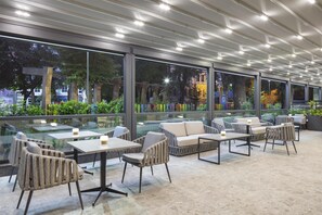 A modern outdoor seating area with tables and chairs, large windows, and a view of the cityscape. - Doubletree By Hilton Plovdiv Center (Plovdiv)