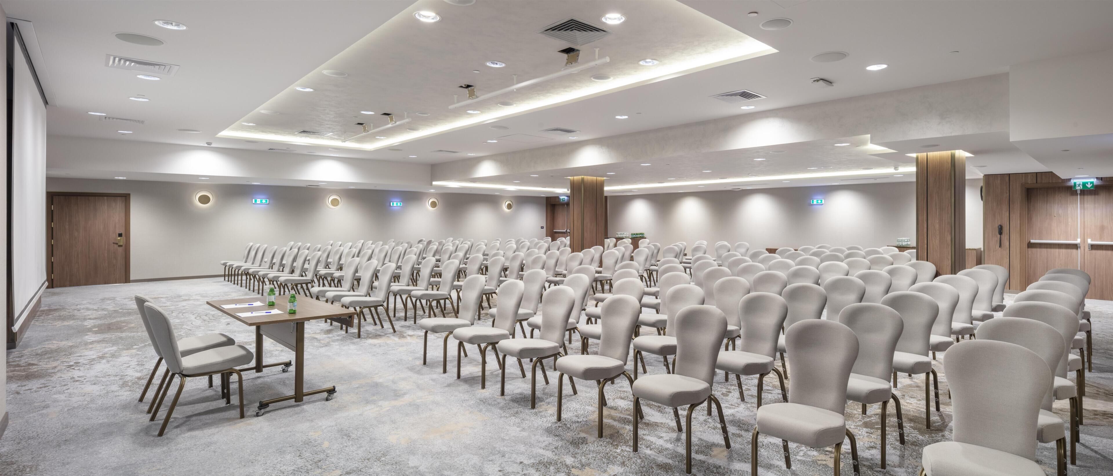 Un amplio salón de conferencias con filas de sillas, mesas y suelo alfombrado.