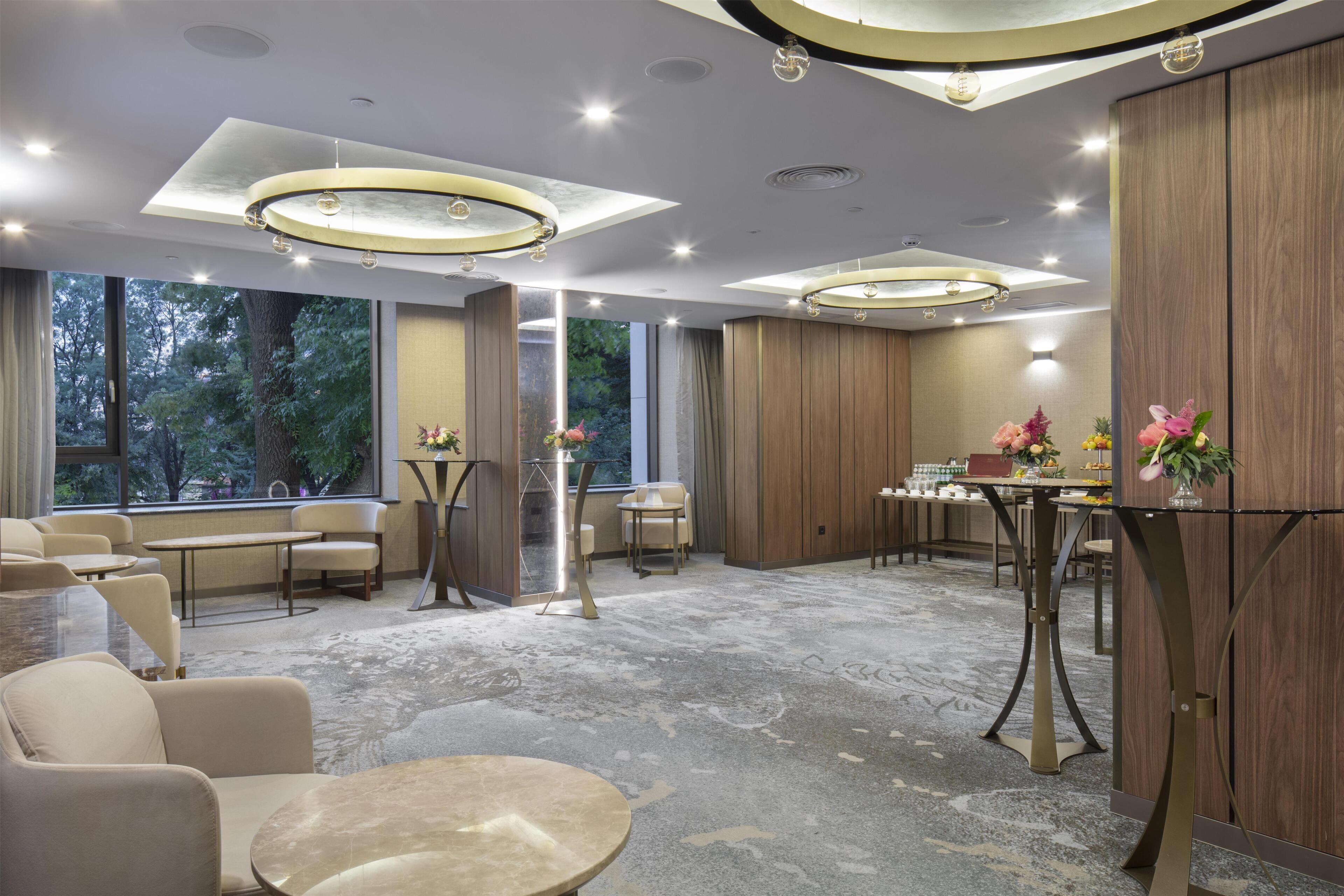 Een moderne hotellobby met ronde tafels, zachte zitplaatsen, een centrale pilaar en grote ramen met uitzicht op bomen.