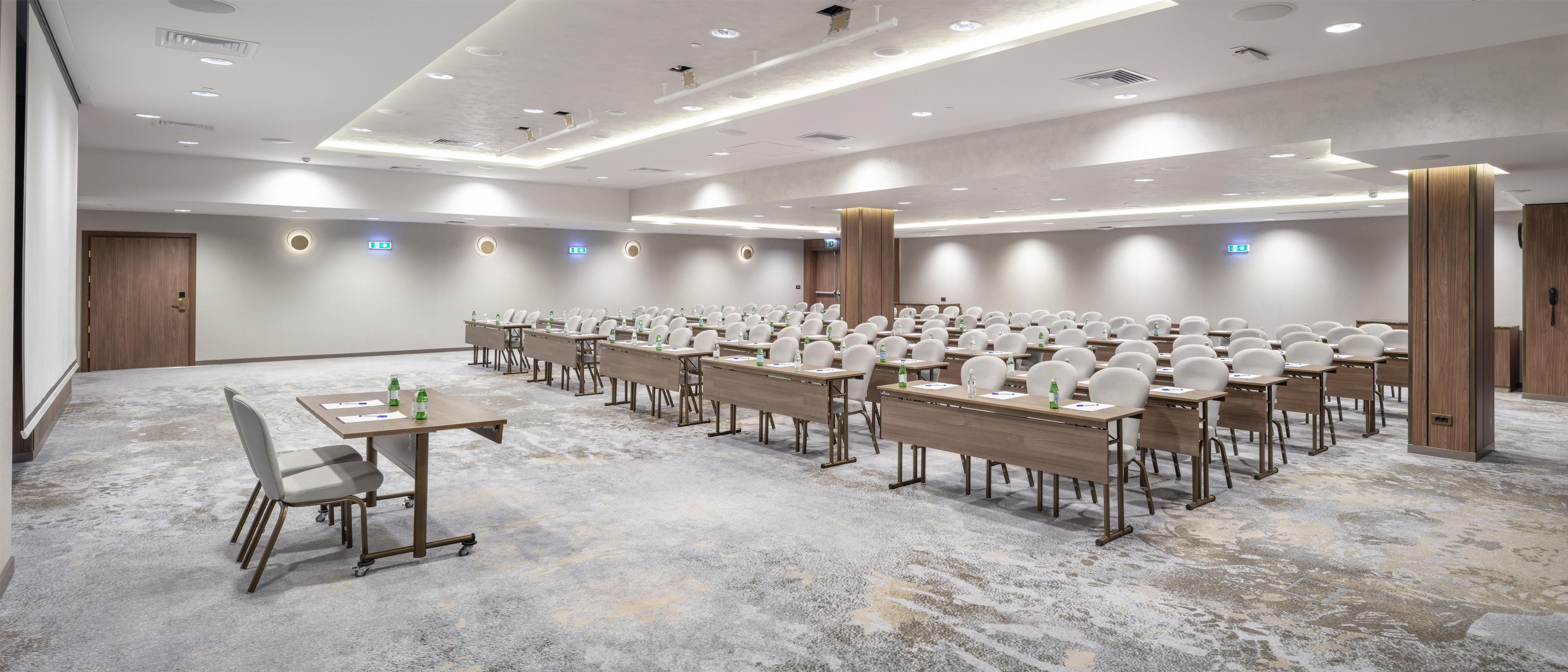 Un salón de conferencias con filas de mesas y sillas, equipado con botellas de agua y documentos, y piso alfombrado.