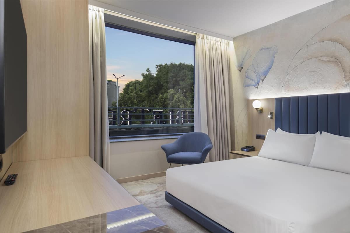 A hotel room with a bed, a chair, a nightstand, a lamp, and a view of trees through a window.