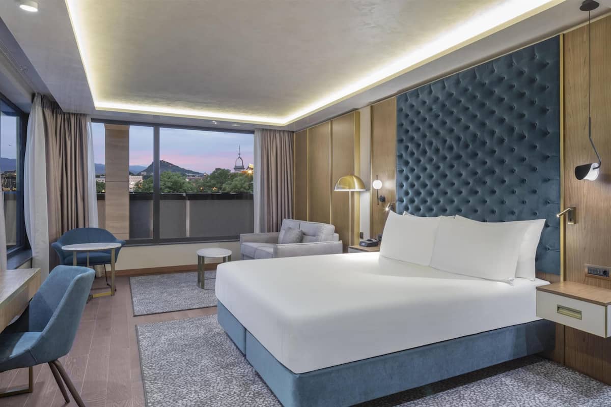 A hotel room with a large bed, a desk, a chair, a small table, a sofa, a TV, and a view of the cityscape.