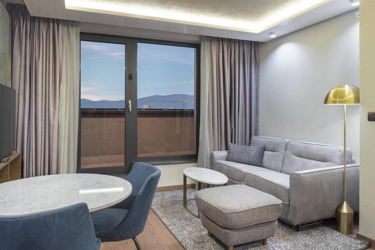 A modern hotel room with a balcony, a sofa, a dining table, and a TV.