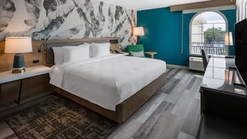 A modern hotel room with a large bed, a desk, a chair, a TV, and a balcony view.