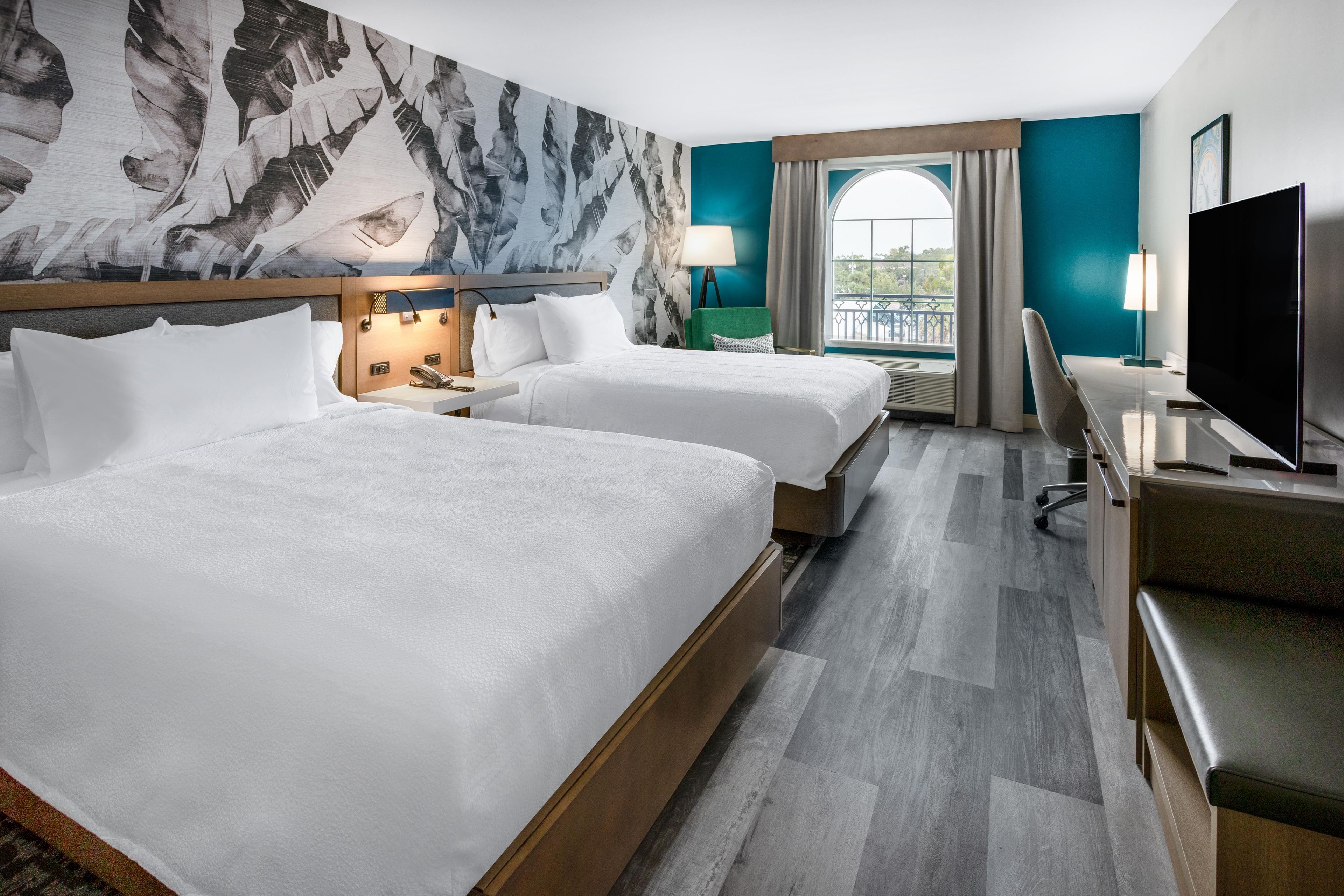 Habitación de hotel con dos camas, un escritorio, televisión y un ventanal con cortinas.