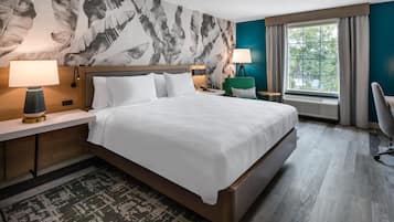 A hotel room with a large bed, two bedside tables with lamps, a green armchair, a desk with a chair, and a window with curtains.