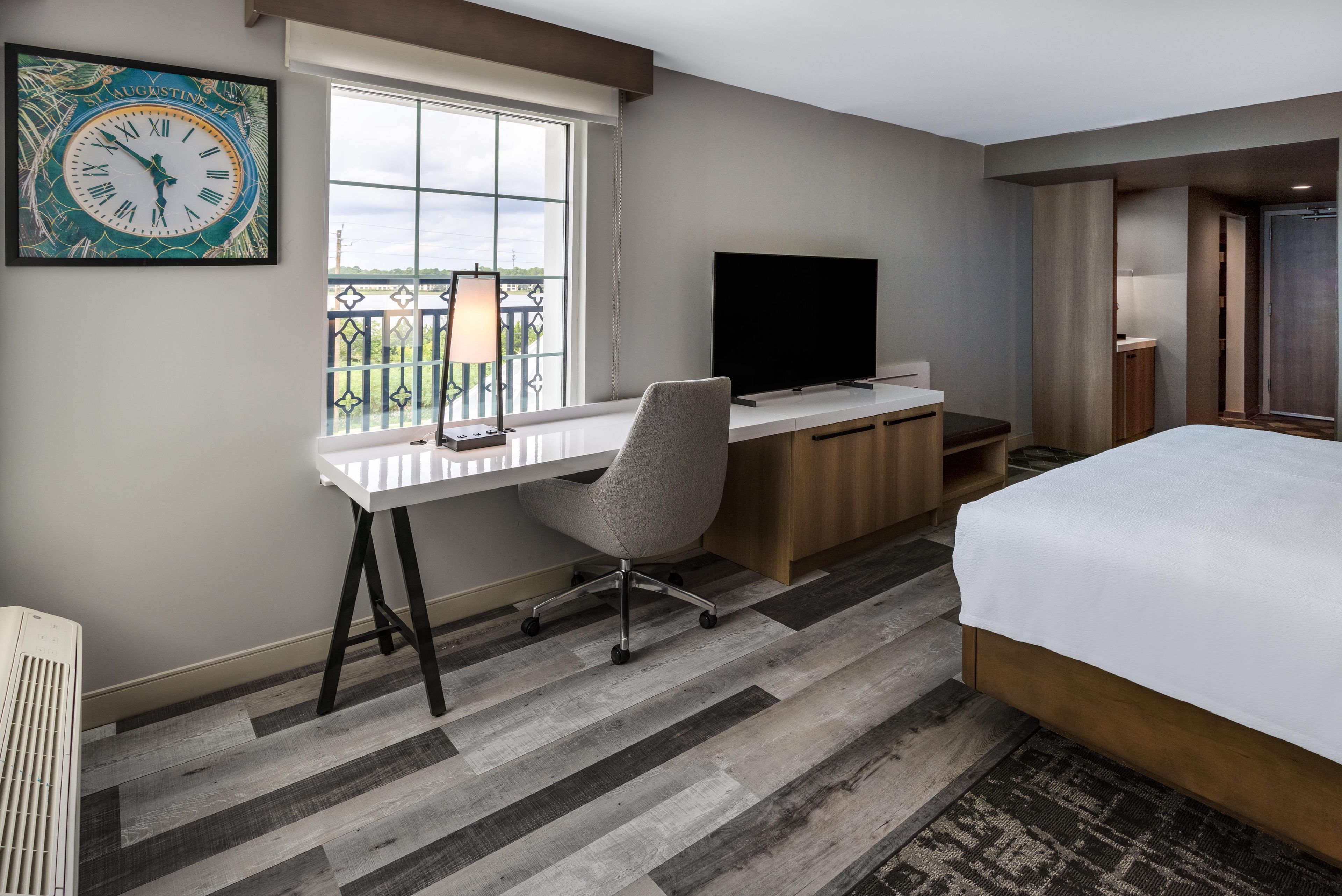A hotel room with a desk, chair, television, bed, and a view of the outdoors.