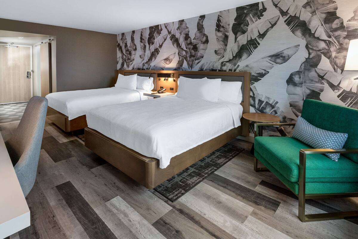 A hotel room with a large bed, a smaller bed, a green armchair, a desk, and a patterned wall.