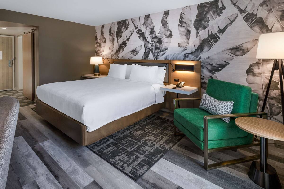 A hotel room with a large bed, a green armchair, a small table, and a patterned wall.