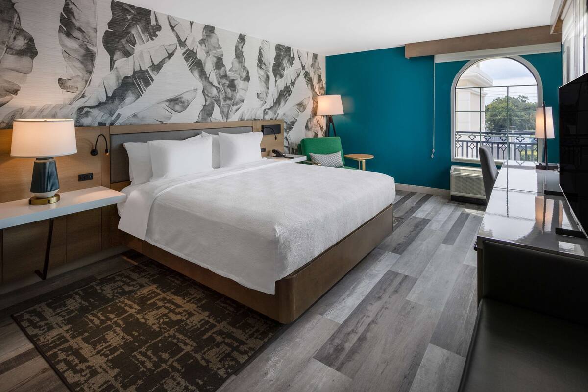 A modern hotel room with a large bed, a desk, a chair, a TV, and a balcony with a view.
