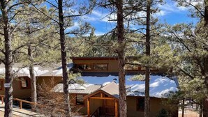 A cabin with a wooden porch and a covered entryway, surrounded by trees and snow. - Walk to ski & trails! Luxury lodge, sleeps 17, hot tub, fire pit & fenced yard. (Angel Fire)