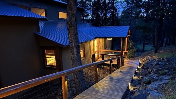 A wooden deck leading to a building with a blue roof, surrounded by trees. - Walk to ski & trails! Luxury lodge, sleeps 17, hot tub, fire pit & fenced yard. (Angel Fire)