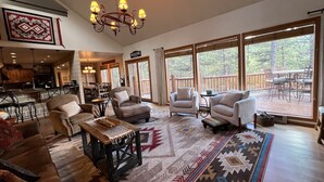 A spacious living room with a large chandelier, comfortable seating, a coffee table, and a view of the outdoors through large windows. - Walk to ski & trails! Luxury lodge, sleeps 17, hot tub, fire pit & fenced yard. (Angel Fire)