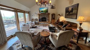 A spacious living room with a fireplace, comfortable seating, and a large window overlooking a deck. - Walk to ski & trails! Luxury lodge, sleeps 17, hot tub, fire pit & fenced yard. (Angel Fire)