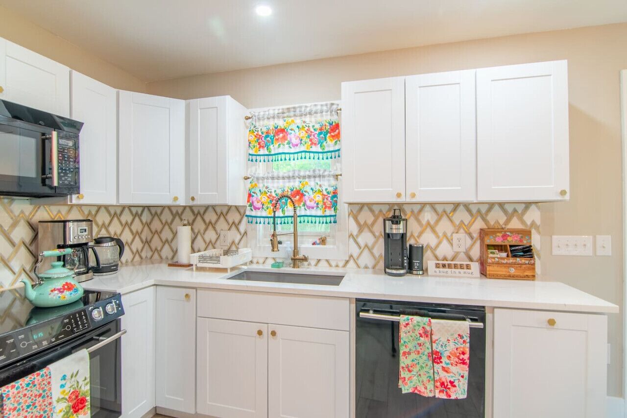 Una cocina con armarios blancos, microondas, estufa, cafetera y ventana con cortinas florales.