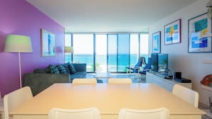A modern living room with a large table, white chairs, a sofa, and a television. There are large windows offering a view of the ocean. - Capri by the Sea 203 | Ocean View | Full Kitchen | Private Bedroom | Beach Access | On the Boardwalk (San Diego)