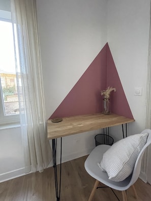A corner desk with a triangular backrest, a chair, a vase with dried flowers, and a window with curtains.