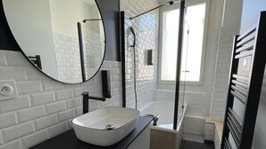 A modern bathroom with a white sink, black countertop, and a large mirror. There is a shower area with a glass door and a patterned floor. - L'Appartement Bleu - Cosy apartment - Heart of town (Douai)