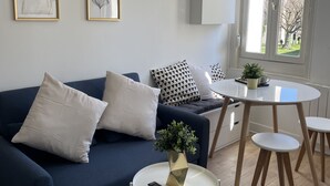 A modern living room with a blue sofa, white cushions, a small round table with a plant, and a window with a view of buildings. - L'Appartement Bleu - Cosy apartment - Heart of town (Douai)