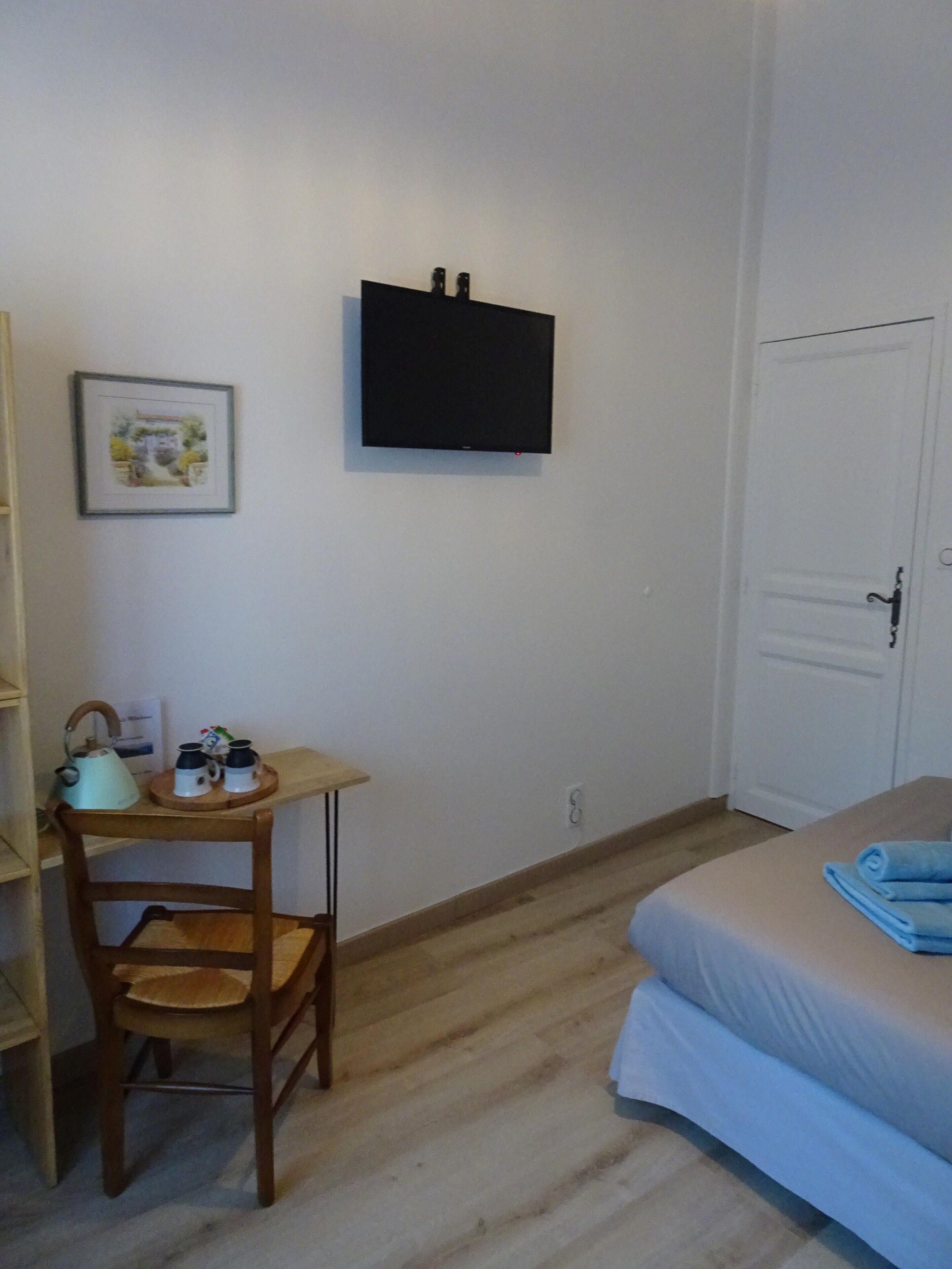 Une petite pièce propre et bien rangée, avec un lit, un bureau et une chaise, une bouilloire et un tableau encadré au mur.